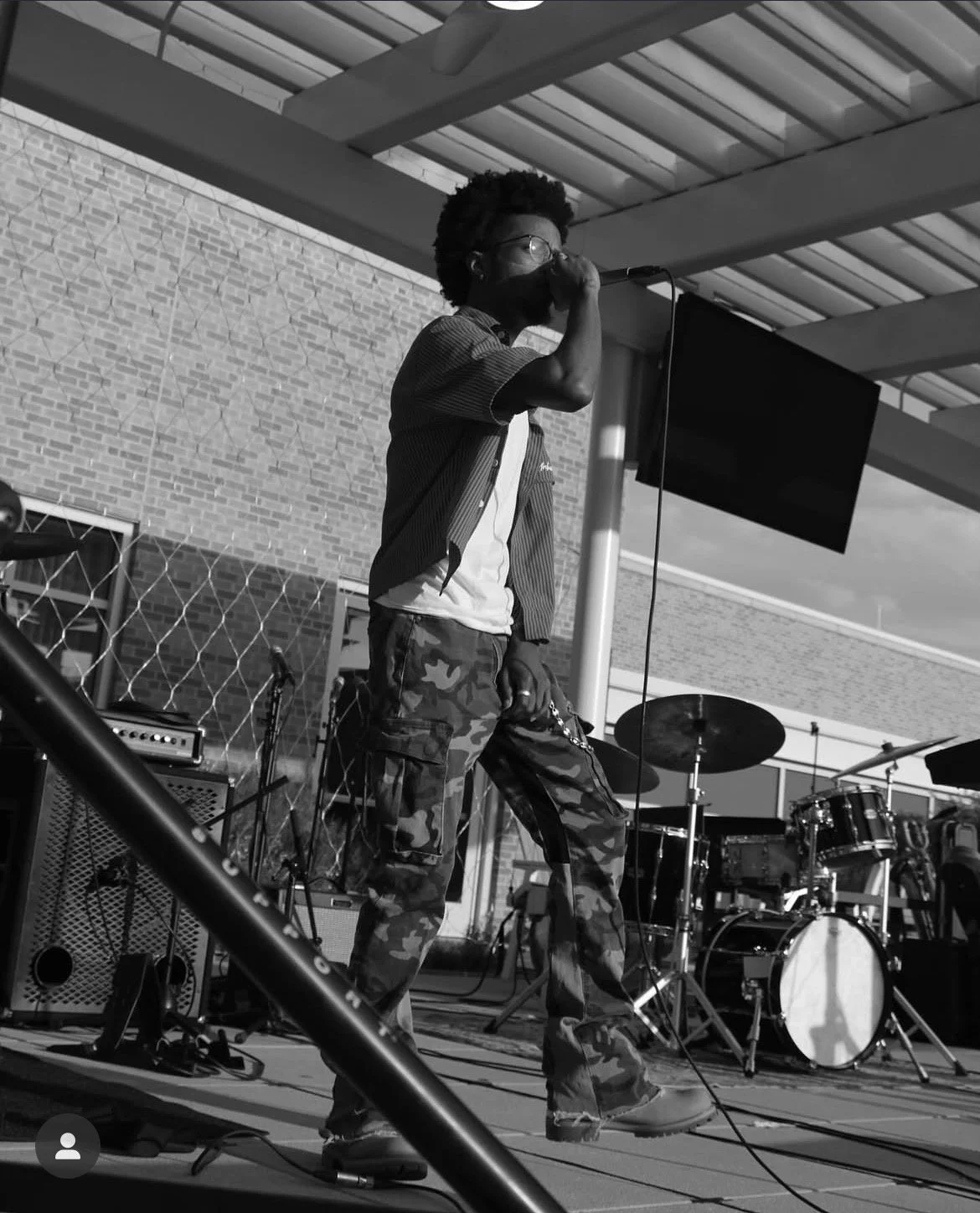 A young man singing into a microphone during an outdoor music performance, with a drum set and amplifier in the background.