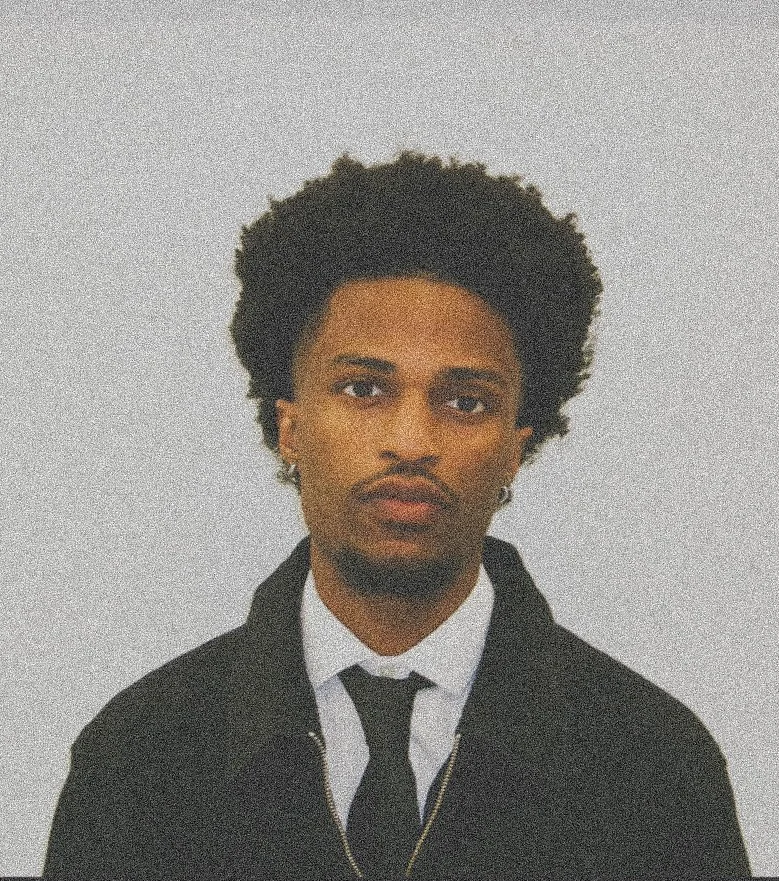 A young man with curly black hair, wearing a black suit, white shirt, black tie, and a gold chain, standing against a plain light background.