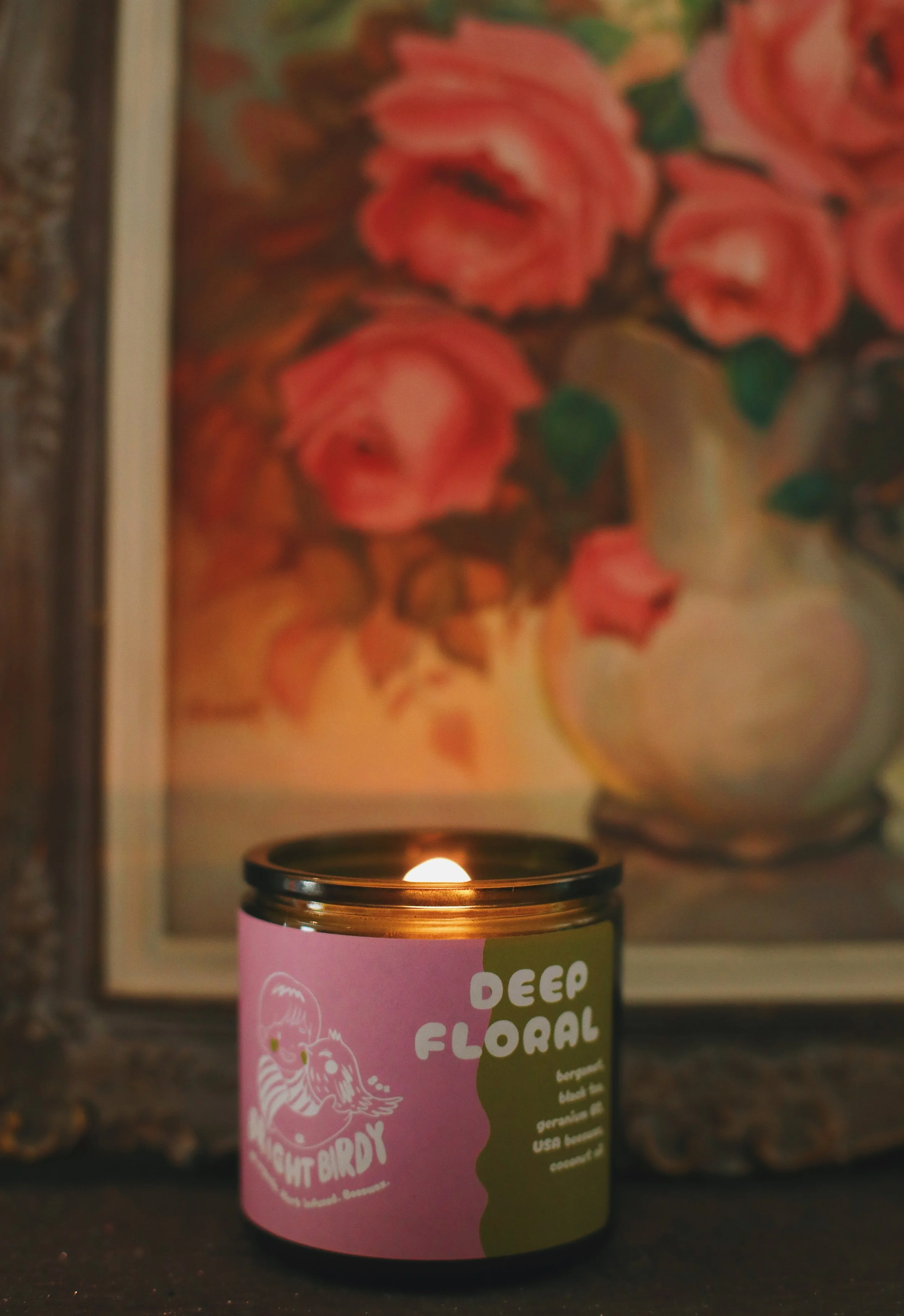 A lit candle in front of a framed painting of pink roses with green leaves. Non-toxic beeswax candle. Moody. 100% organic candles.