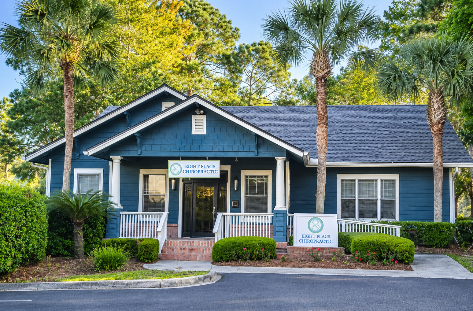 A blue building with white porch railings and columns, with signs for Eight Flags Chiropractic. There are two palm trees in front and lush green bushes with flowers, set in a landscaped area with a curved driveway and sidewalk.