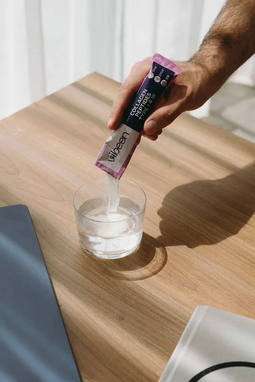 A person pouring a white powder labeled 'Collagen Peptides, Type I & III' into a glass of water on a wooden table.