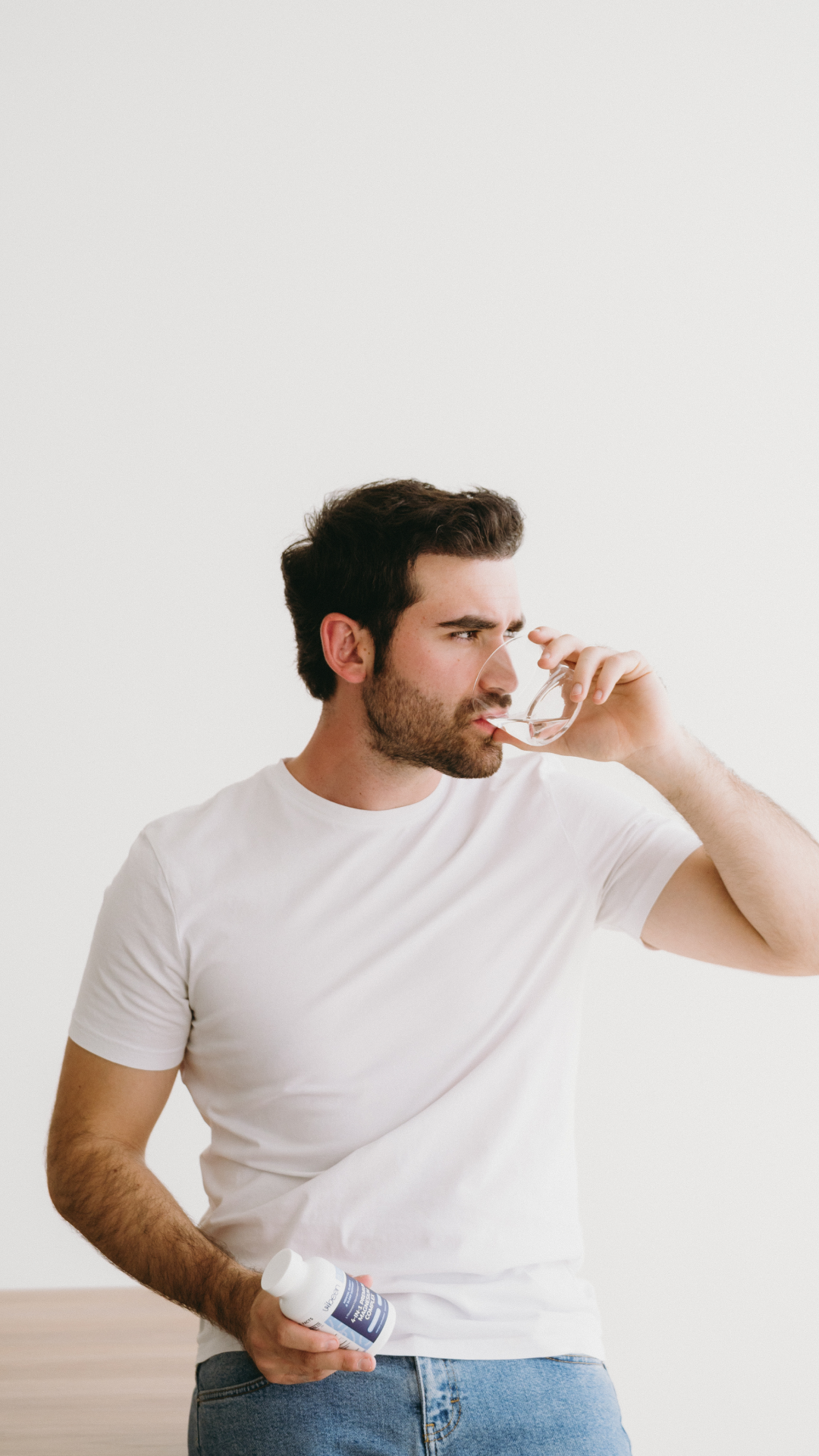 A young man with dark hair and a beard, wearing a white t-shirt and jeans, holding a bottle in his left hand, sniffing a nasal spray with his right hand, standing against a plain white background.