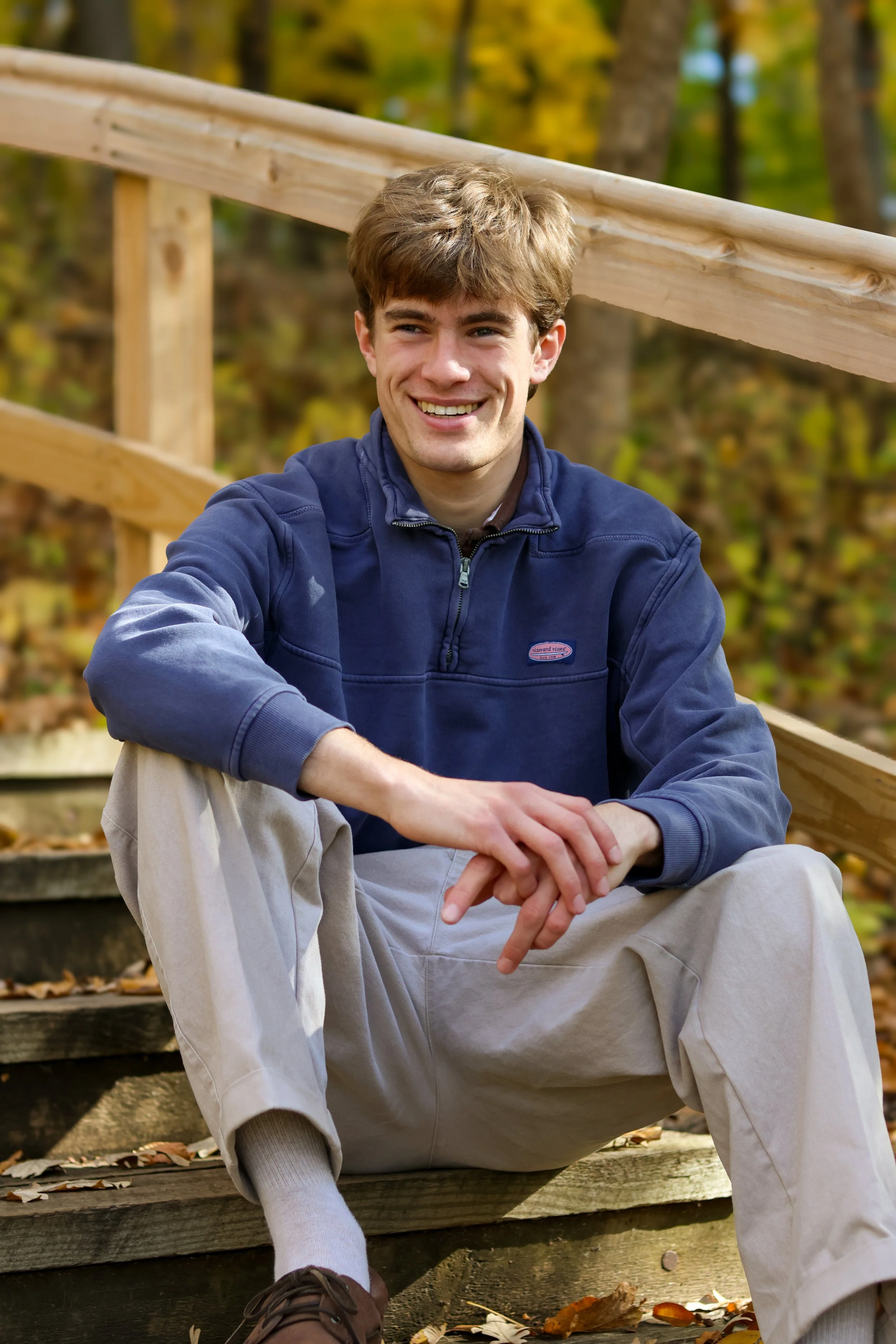 A young man with brown hair smiling while sitting on outdoor wooden steps amidst fall foliage, wearing a blue jacket, beige pants, and brown shoes.