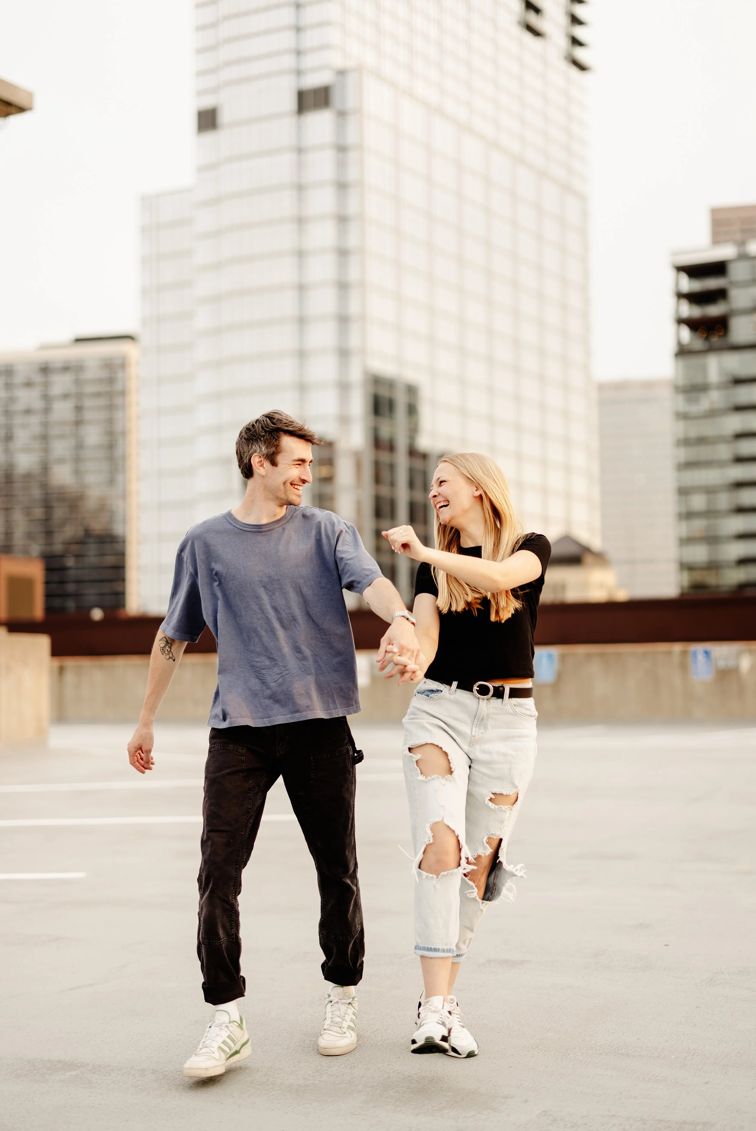 A young man and woman are walking and laughing together in an urban parking lot with tall modern buildings in the background. The woman is wearing ripped jeans and white sneakers, and the man is wearing a gray t-shirt, black jeans, and white sneakers. They are holding hands and seem to be enjoying each other's company.
