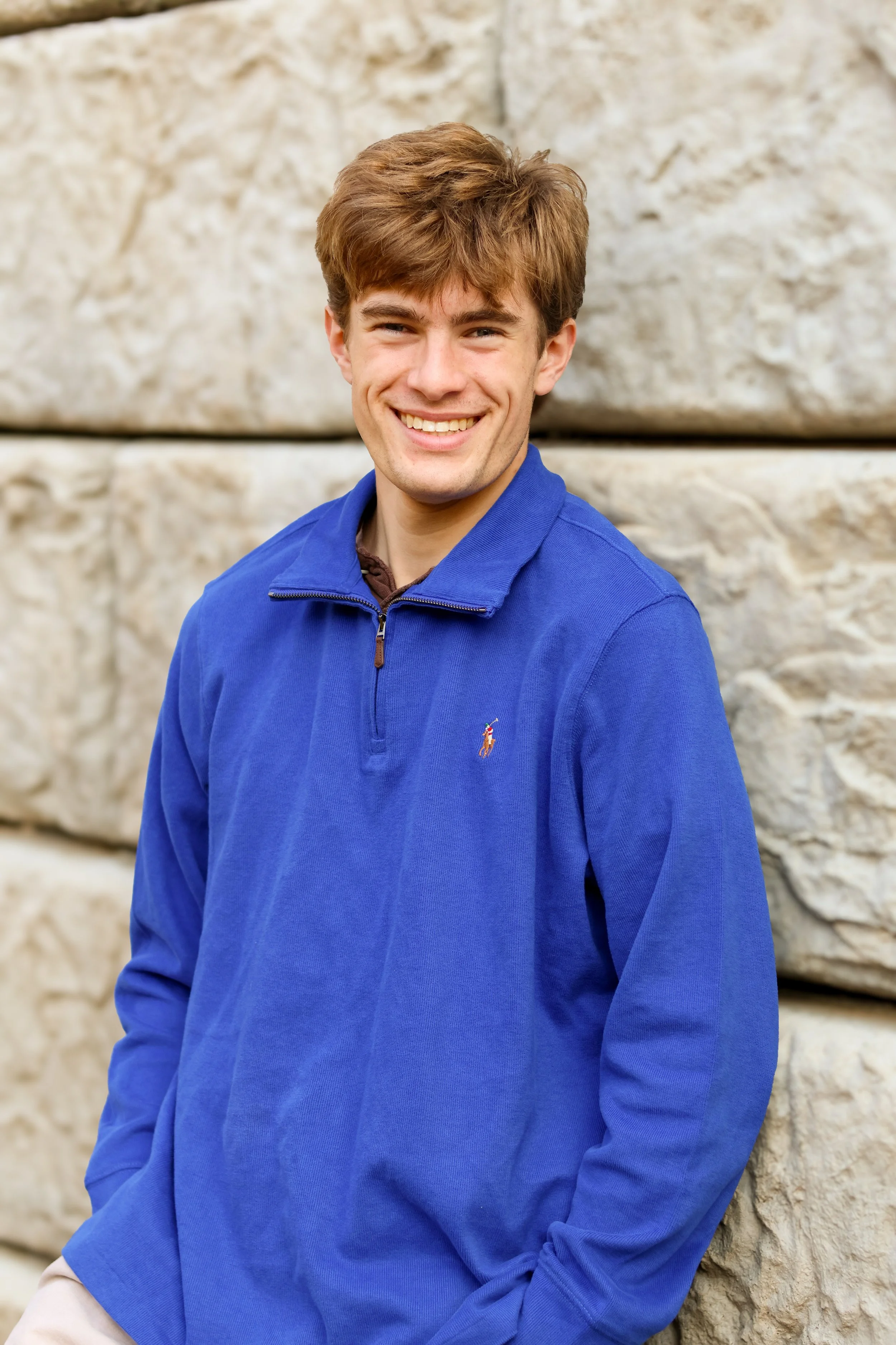 A young man with brown hair smiling, wearing a blue quarter-zip sweater, standing in front of a stone wall.