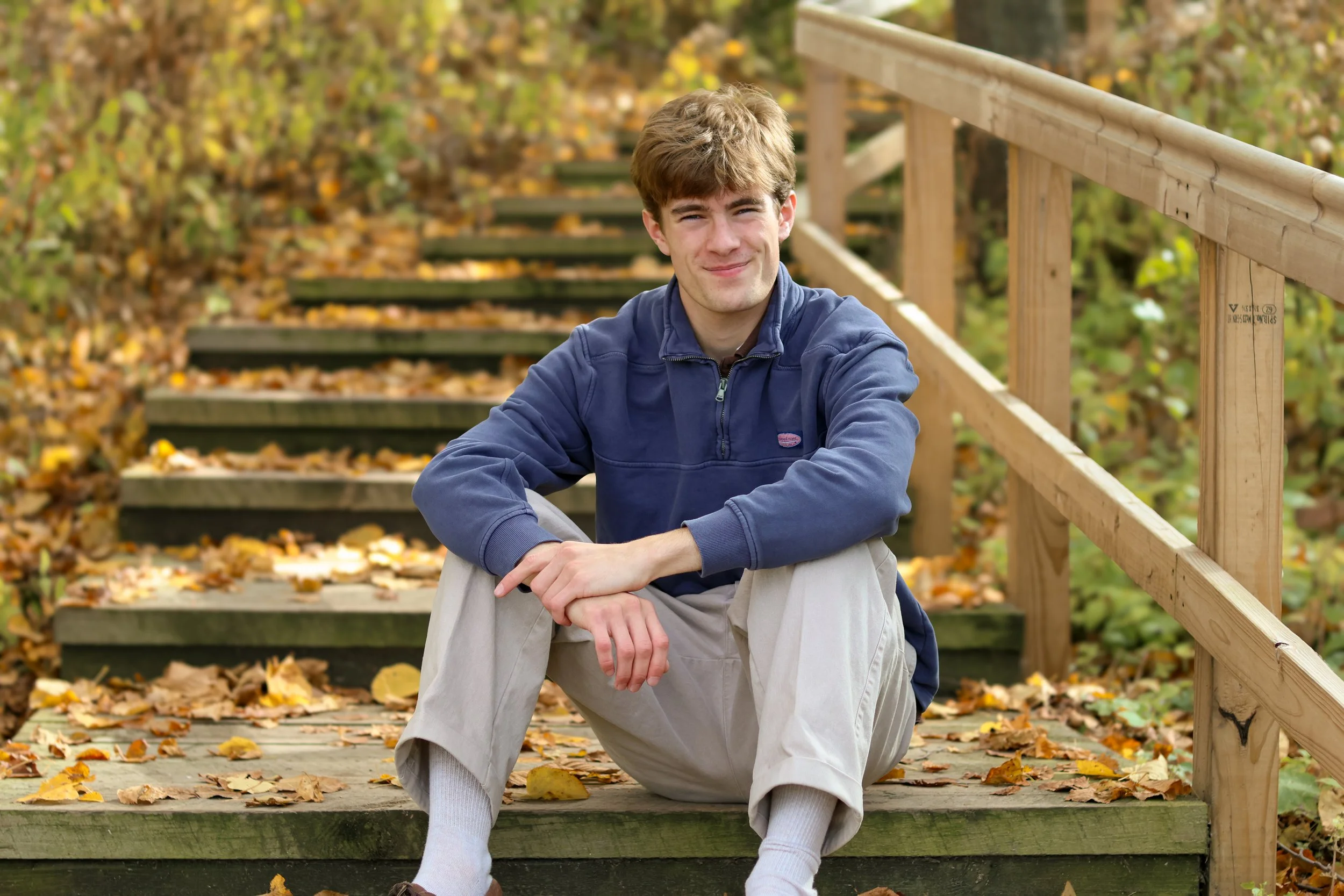A young man with light brown hair and fair skin, wearing a navy blue jacket, beige pants, and white socks, sitting on wooden stairs covered with fallen autumn leaves, smiling at the camera in an outdoor setting with a wooden railing and trees in the background.