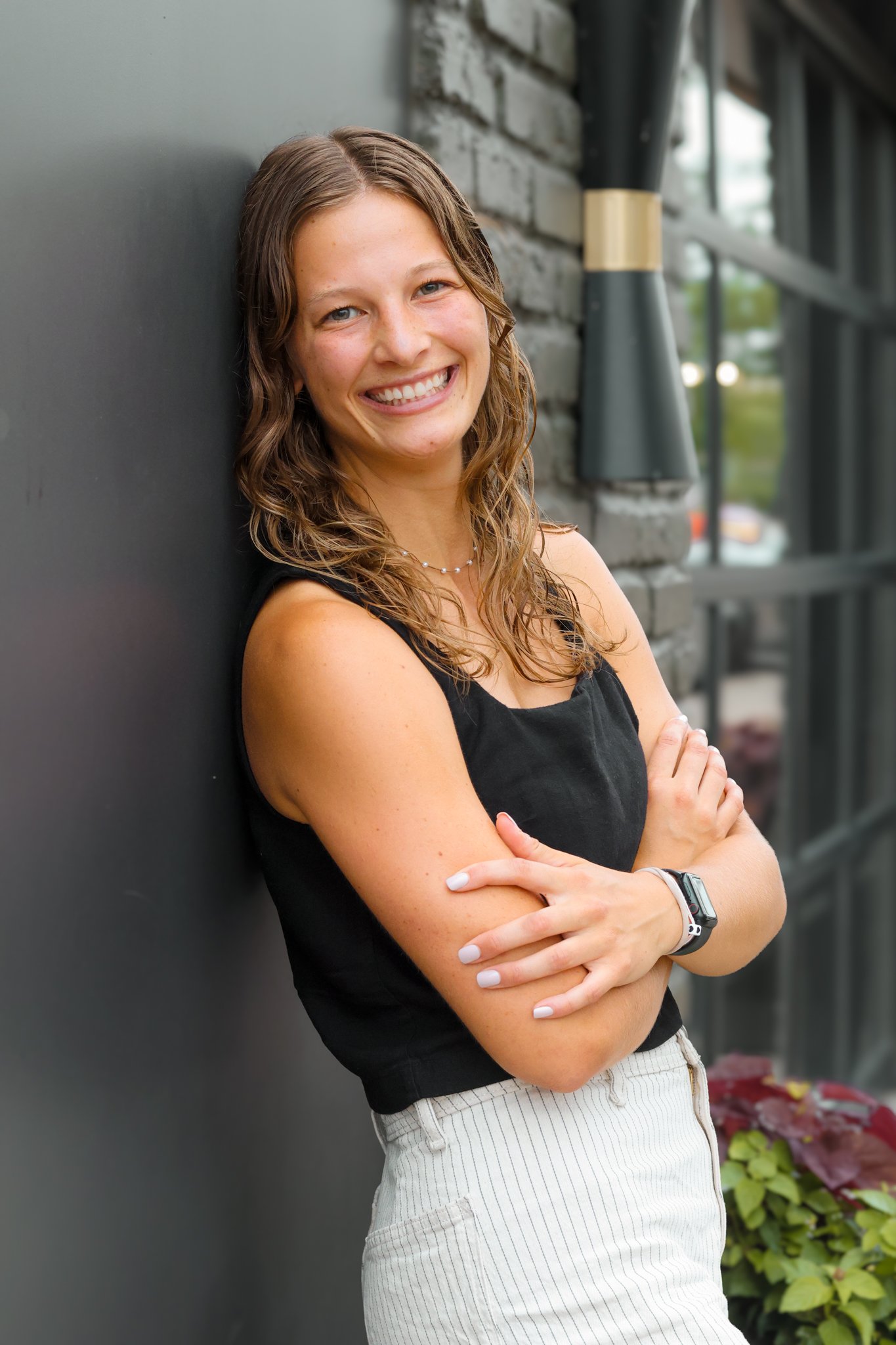 A young woman with wavy brown hair, smiling with arms crossed, standing outdoors against a gray wall with a brick building and windows in the background.
