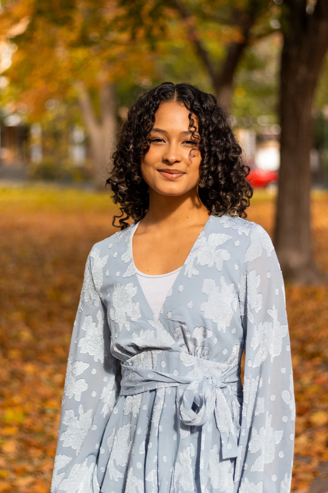 A woman with curly dark hair, wearing a light blue dress with white floral embroidery, standing outdoors in an autumn setting with trees and fallen leaves in the background.