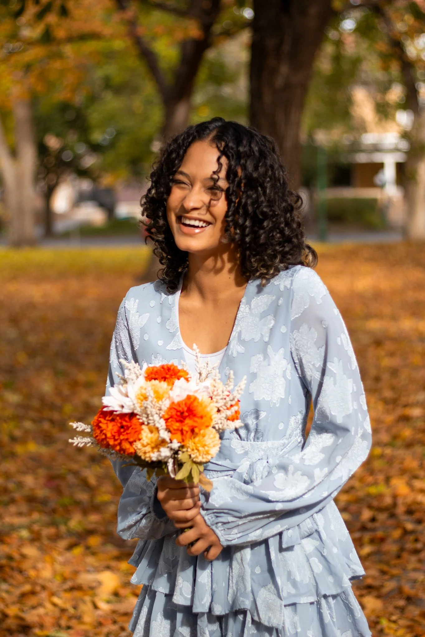 A woman with curly black hair wearing a light blue dress with white floral patterns, holding a bouquet of orange and white flowers, standing outdoors in a park during autumn with fallen leaves on the ground and trees in the background.