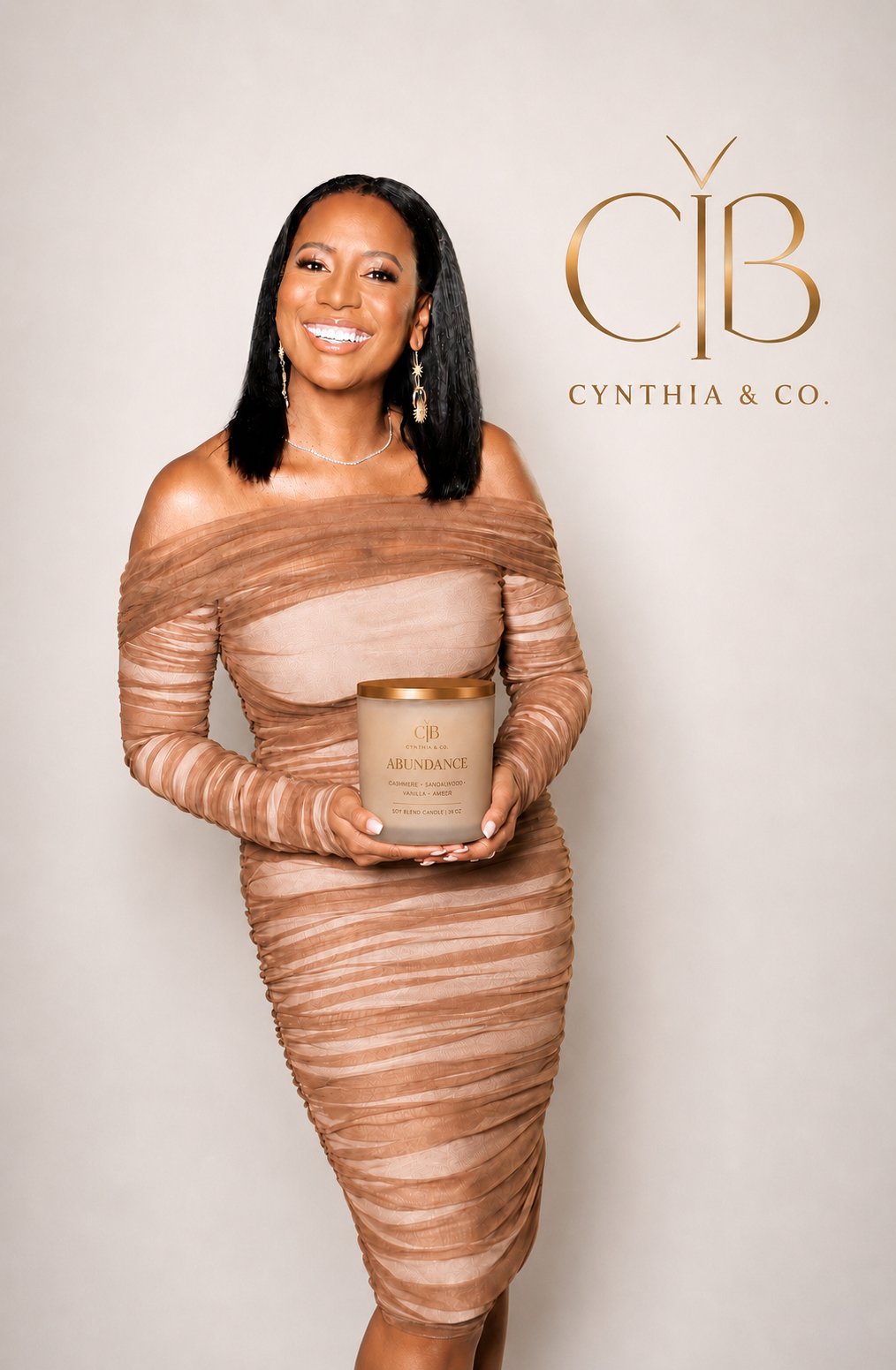 A woman with black hair, dressed in a tan, off-the-shoulder ruched dress, holding a beige candle labeled 'Abundance' with scent notes, standing in front of a white background with a gold Cynthia & Co. logo.