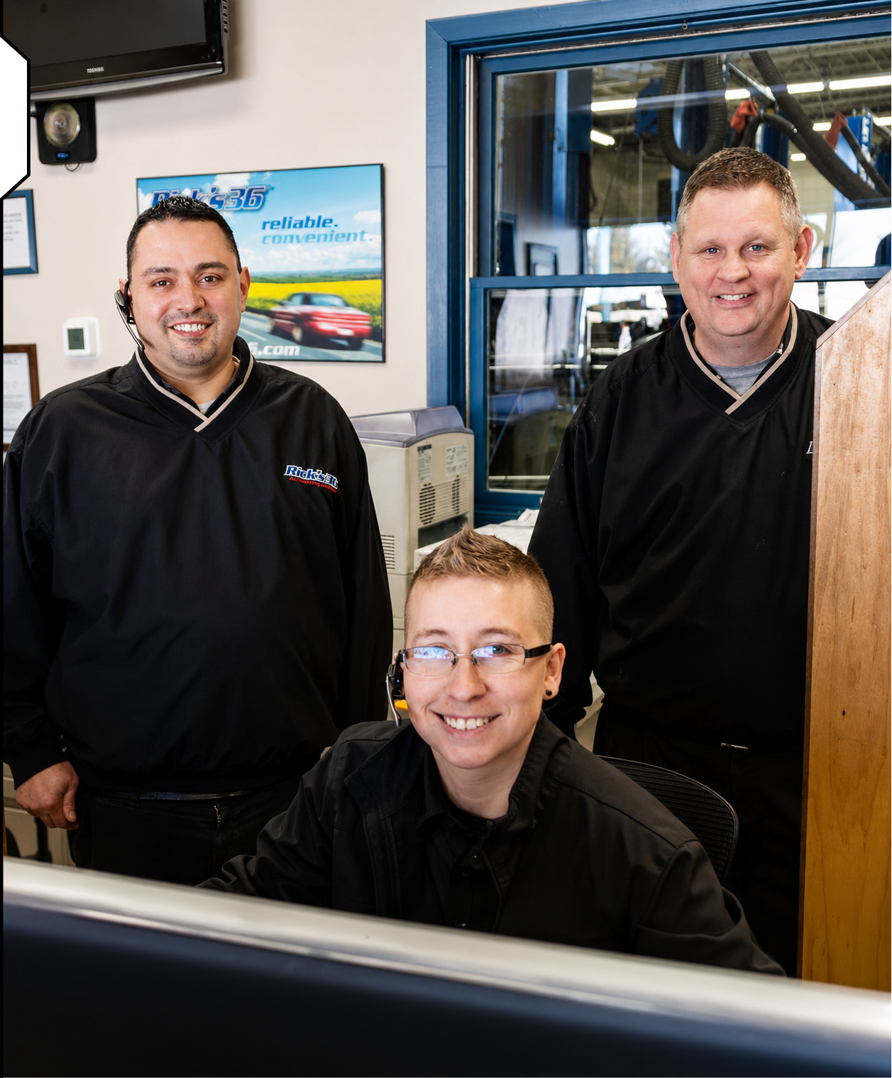 Three employees in a car dealership office. Two standing men wearing black uniforms and one sitting woman with glasses. Behind them are office windows and a display with a car advertisement.
