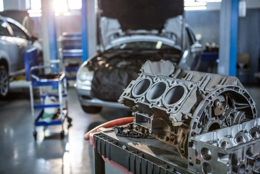Automotive engine block on workbench in auto repair shop with cars and tools in background.