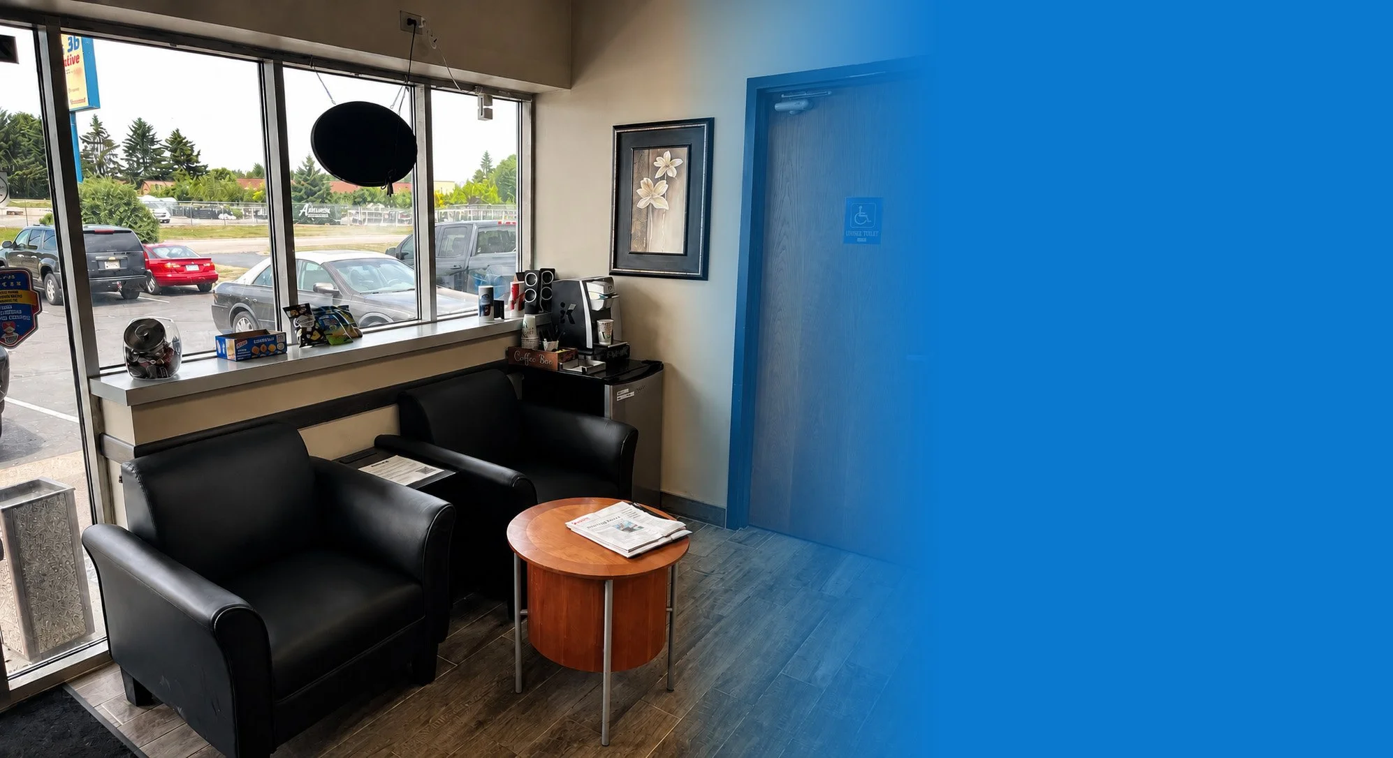 The lobby of an auto shop with black armchair seating, a small wooden table with newspapers, a coffee station, and large windows overlooking parked cars and trees outside.