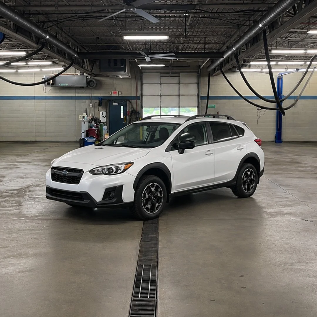 A white Subaru Crosstrek parked inside an automotive shop with equipment and tools in the background.