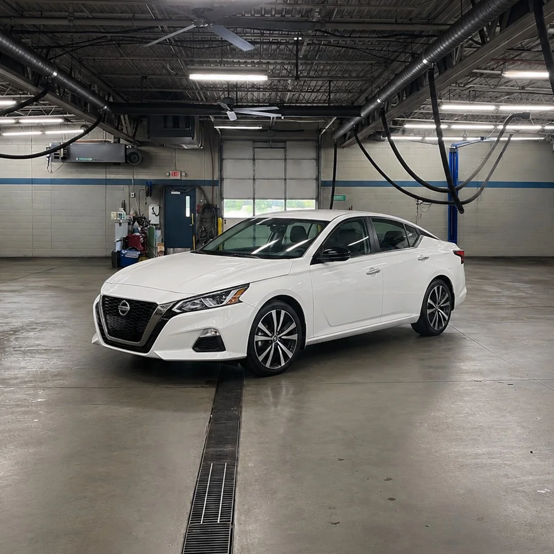 A white Nissan Ultima sedan car parked inside a spacious, industrial-style automotive shop with concrete floors and exposed ceiling pipes and ducts.