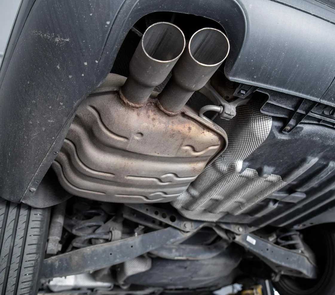 The underside of a vehicle showing the exhaust system with dual exhaust pipes and a rusty catalytic converter.