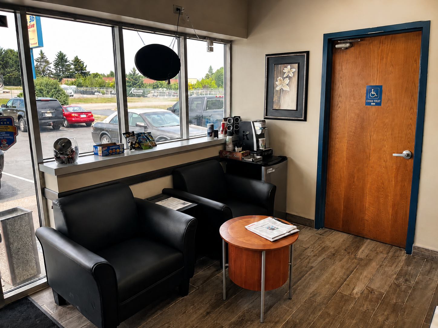 Lobby of an auto-shop with black leather armchairs, a round wooden table with newspapers, a coffee station with cups, a coffee maker, and dispenser, and a framed flower picture. Large window showing a parking lot outside.