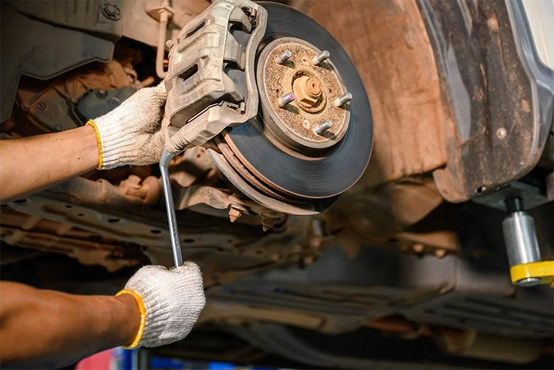 A mechanic wearing gloves is working on a car's brake disc and caliper assembly, which is lifted off the ground in a garage.