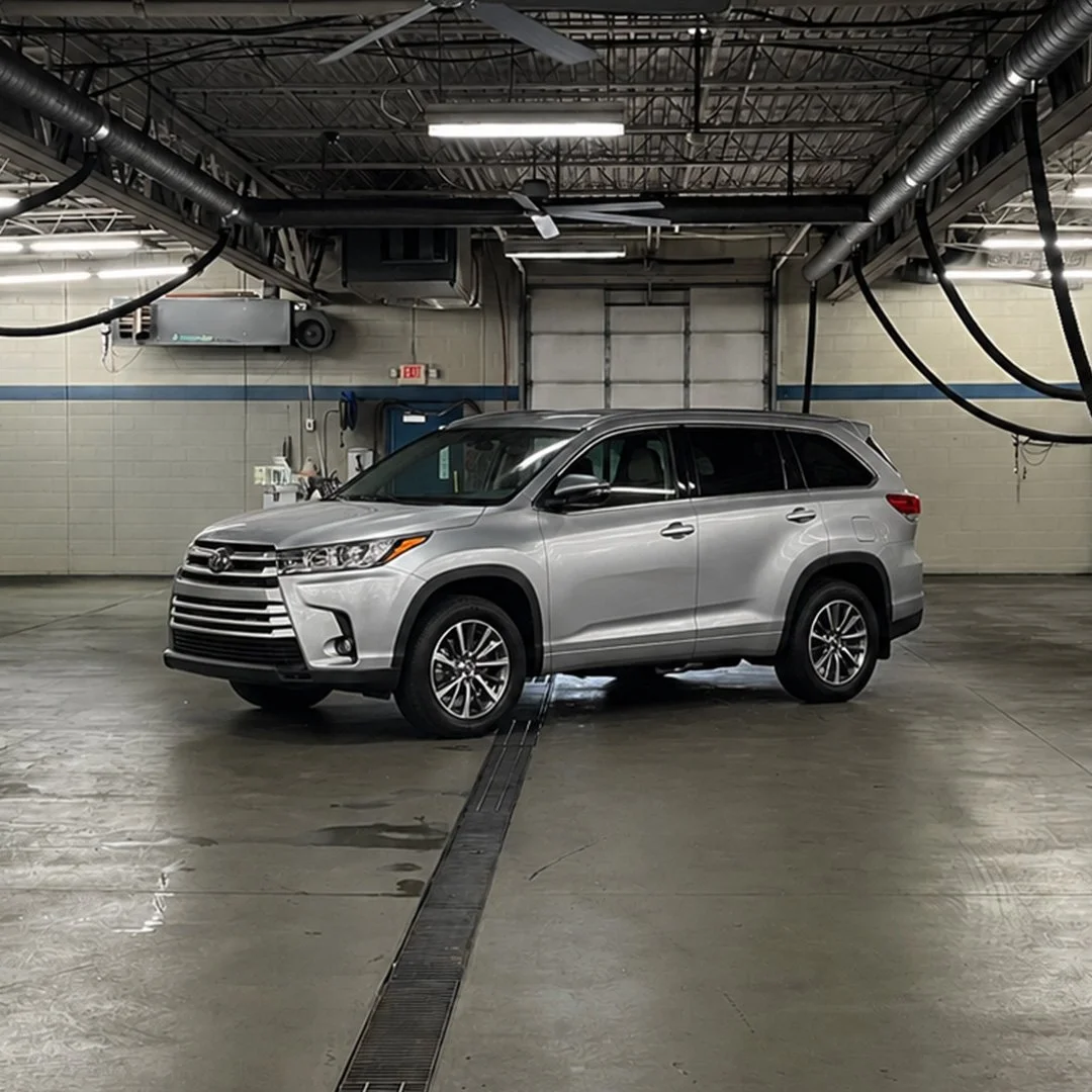 Silver Toyota Highlander SUV parked inside an automotive repair shop with overhead pipes and bright fluorescent lighting.