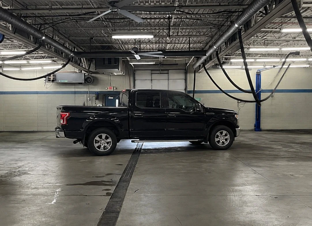 A black Ford F-150 pickup truck parked inside an empty, auto-shop garage or repair shop with exposed ceiling pipes and fluorescent lighting.