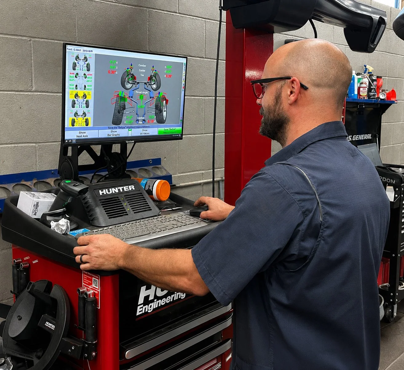 A man with glasses and a beard wearing a navy shirt working at an automotive computer station with a monitor displaying wheel alignment data, car parts, and tools nearby.
