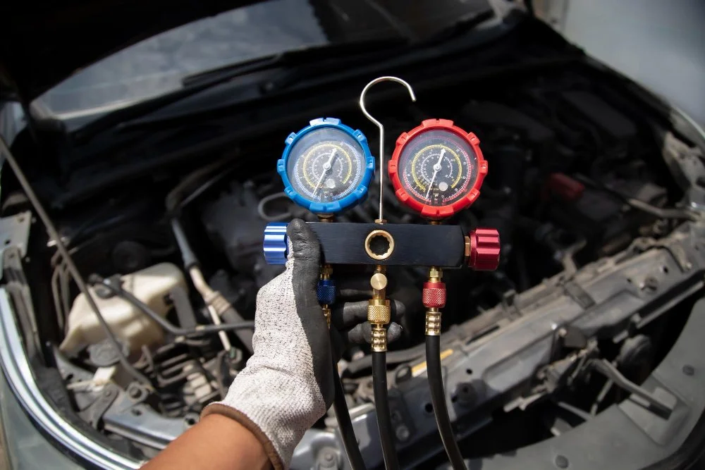A person wearing gloves holding a manifold gauge set with red and blue dials, connected to a car's open engine bay.