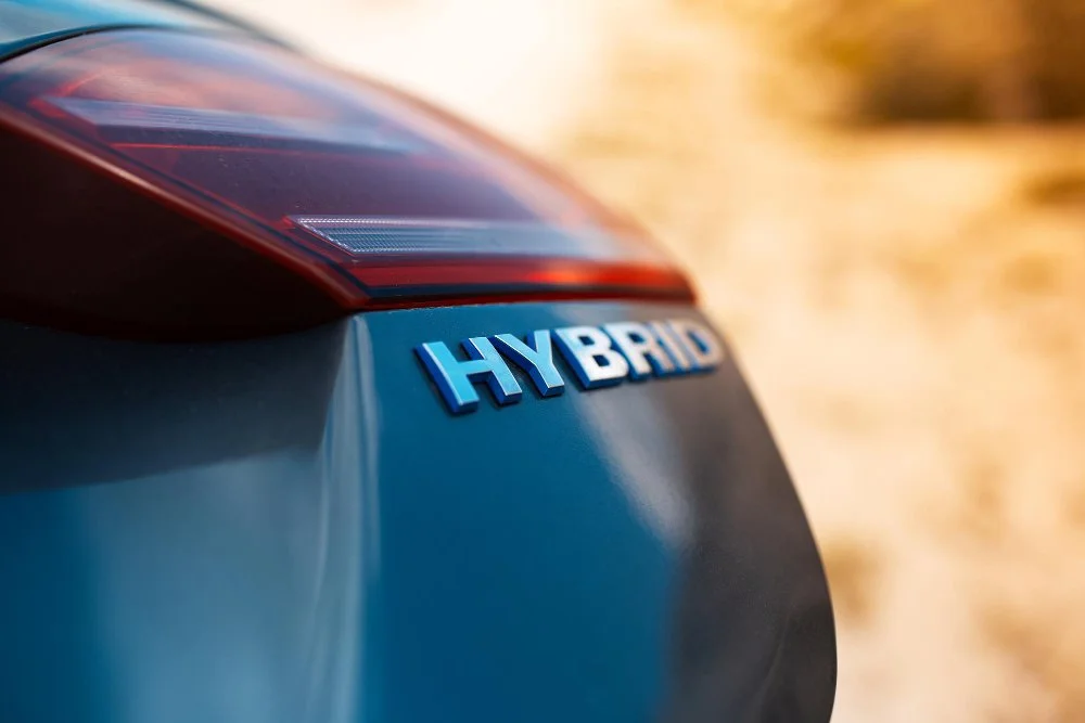 Close-up of the rear of a hybrid car, showing a blue "HYBRID" badge and part of the taillight, with a blurred background in sunlight.
