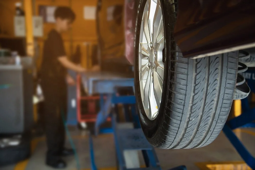 Close-up of a car wheel and tire being serviced in an auto repair shop, with a mechanic working in the background.