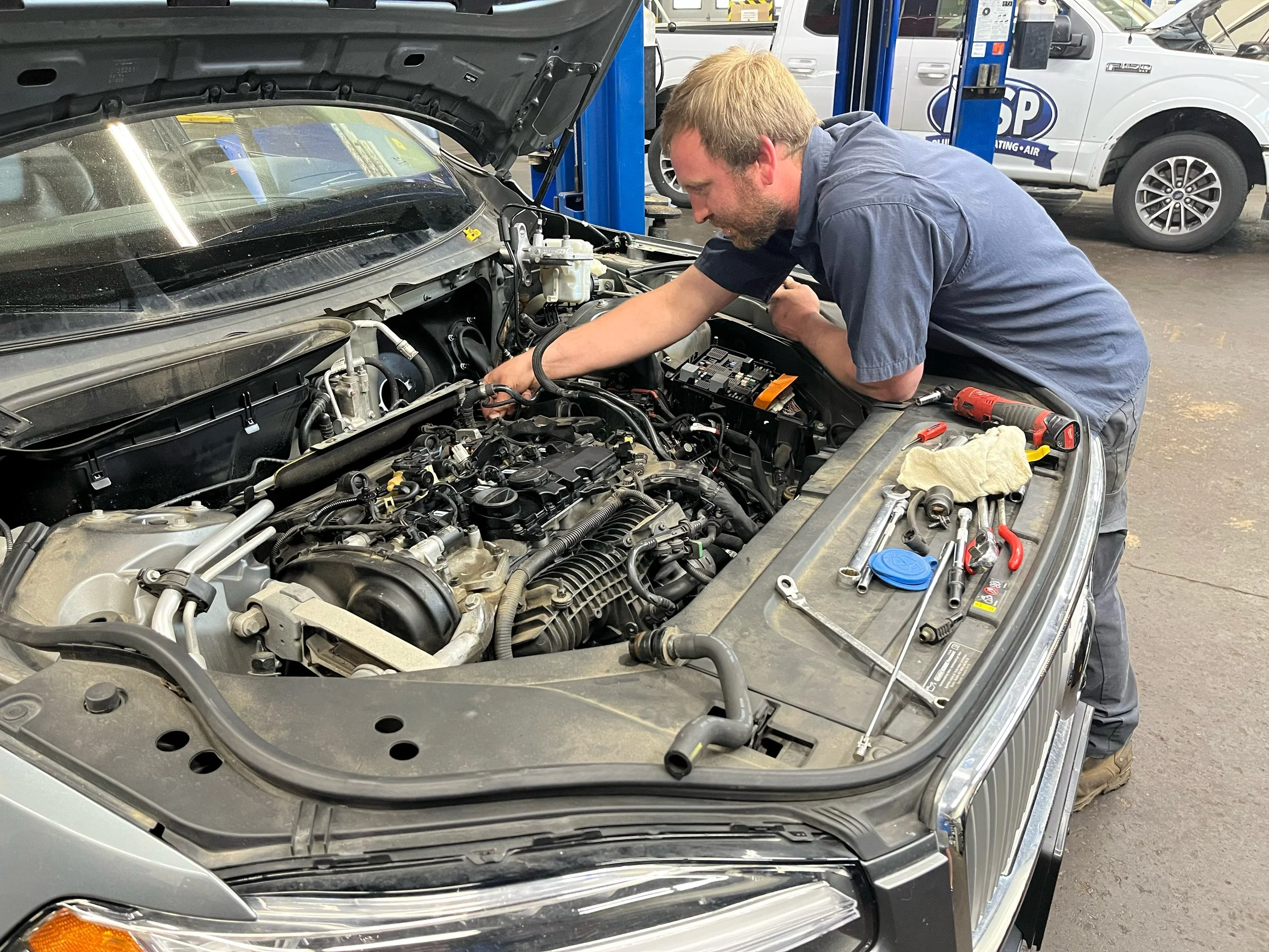 A man working under the hood of a silver car in an auto repair shop, surrounded by tools and equipment, with other vehicles visible in the background.