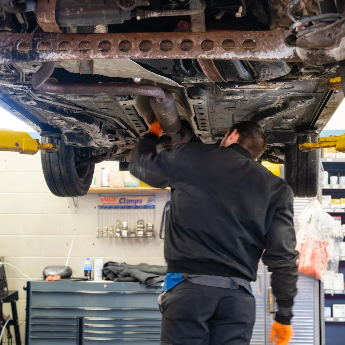 A mechanic working underneath a lifted car in an auto repair shop, wearing orange gloves and a black jacket.