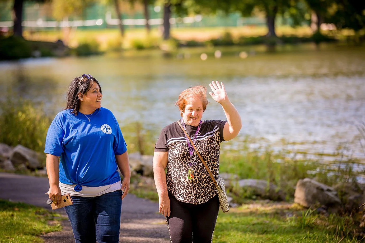 Two smiling individuals walking outside by pond on a sunny day.   One individual is waving