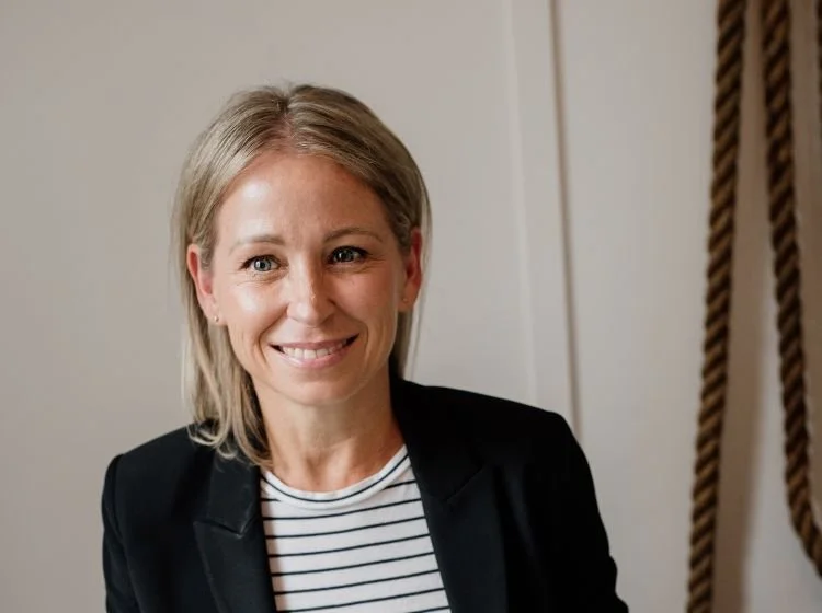 A woman with shoulder-length blonde hair, wearing a black blazer over a striped shirt, smiling at the camera indoors.