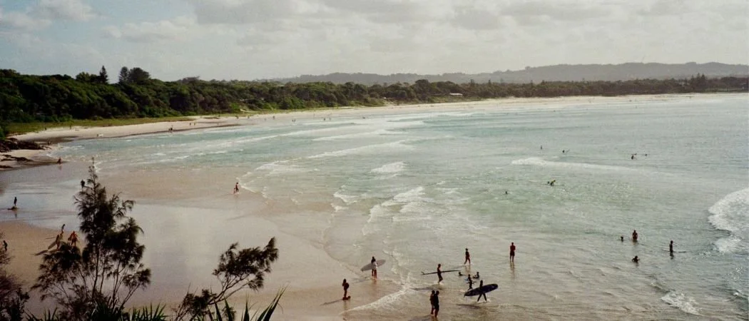 People surfing and swimming at a sandy beach with green trees in the background and cloudy skies.