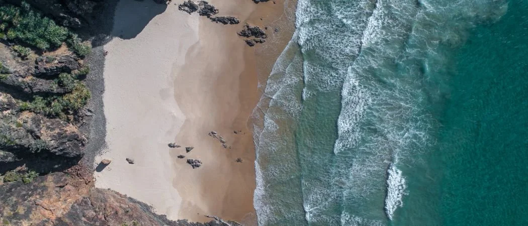aerial view of a beach with sand, waves, and rocky cliffs on one side.