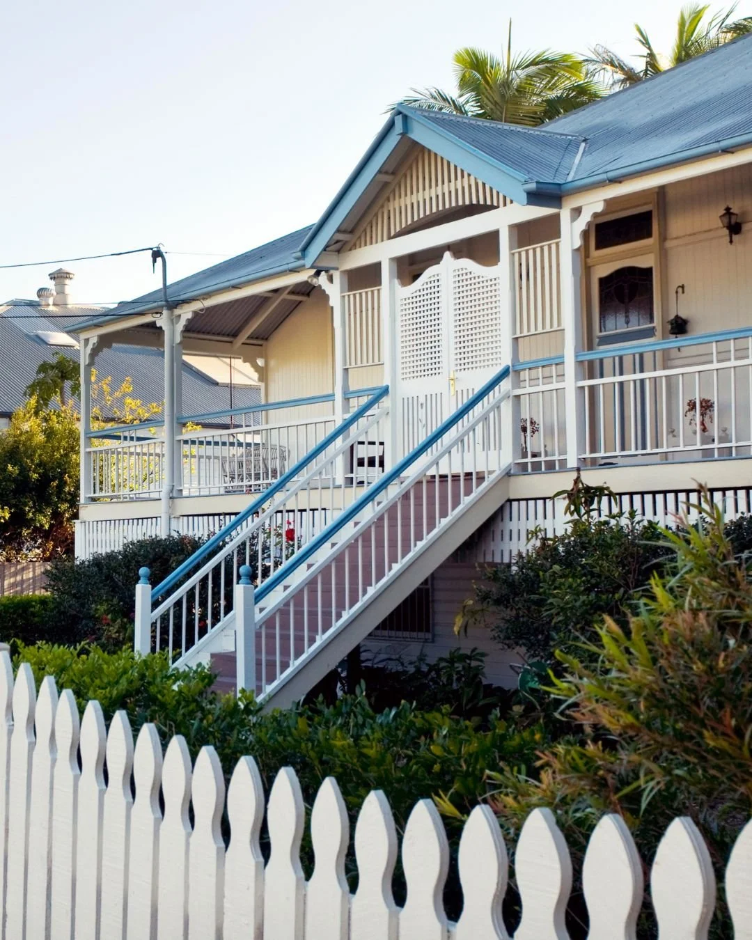 A two-story house with a staircase leading to the front porch. The house is painted white with blue accents on the roof and railing, and has a picket fence in the foreground. There are trees and shrubs surrounding the house.