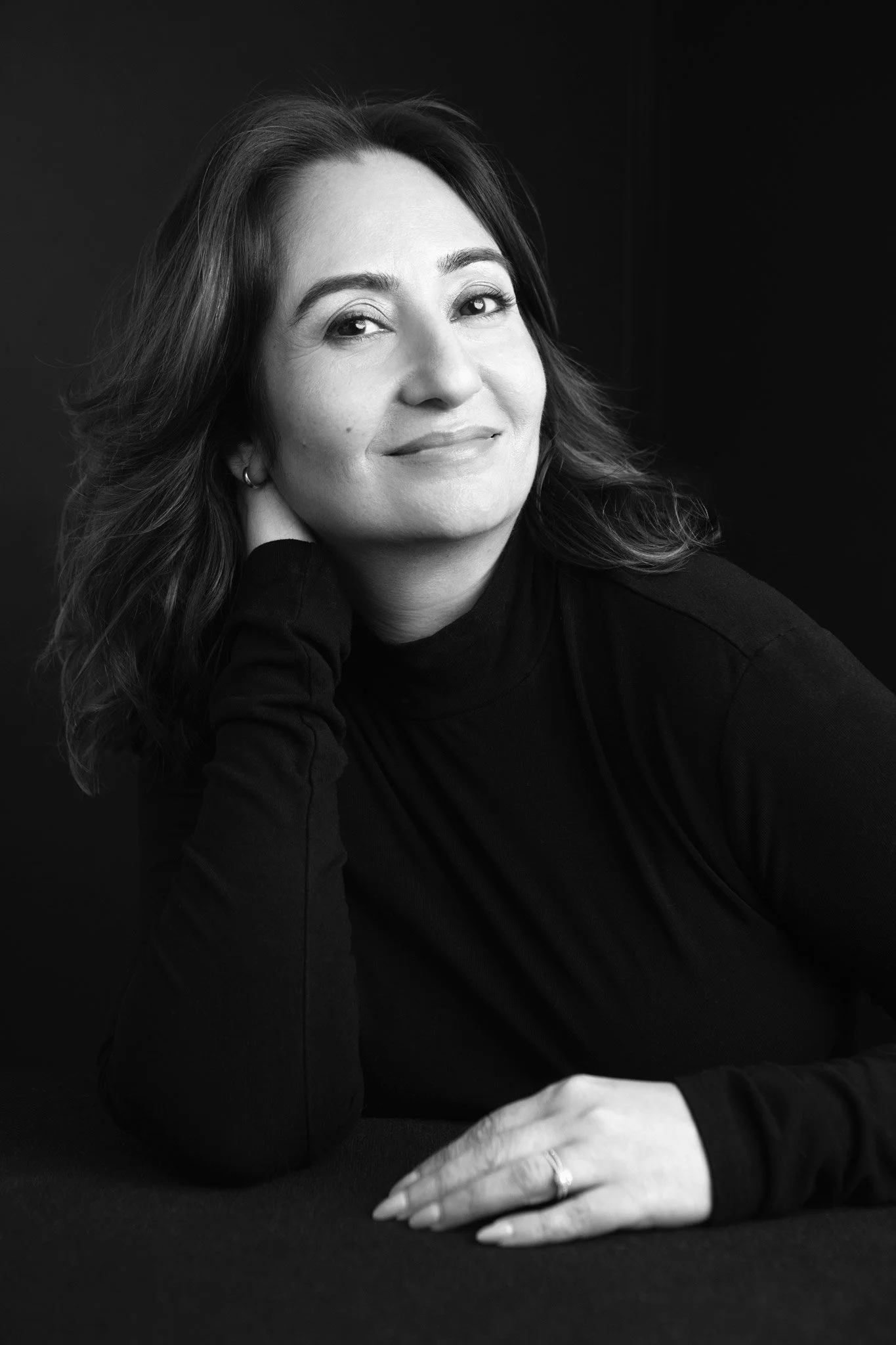 Black and white portrait of a woman with wavy hair, wearing a dark turtleneck, resting her head on her hand, smiling softly, with her hand and a ring visible in the foreground.