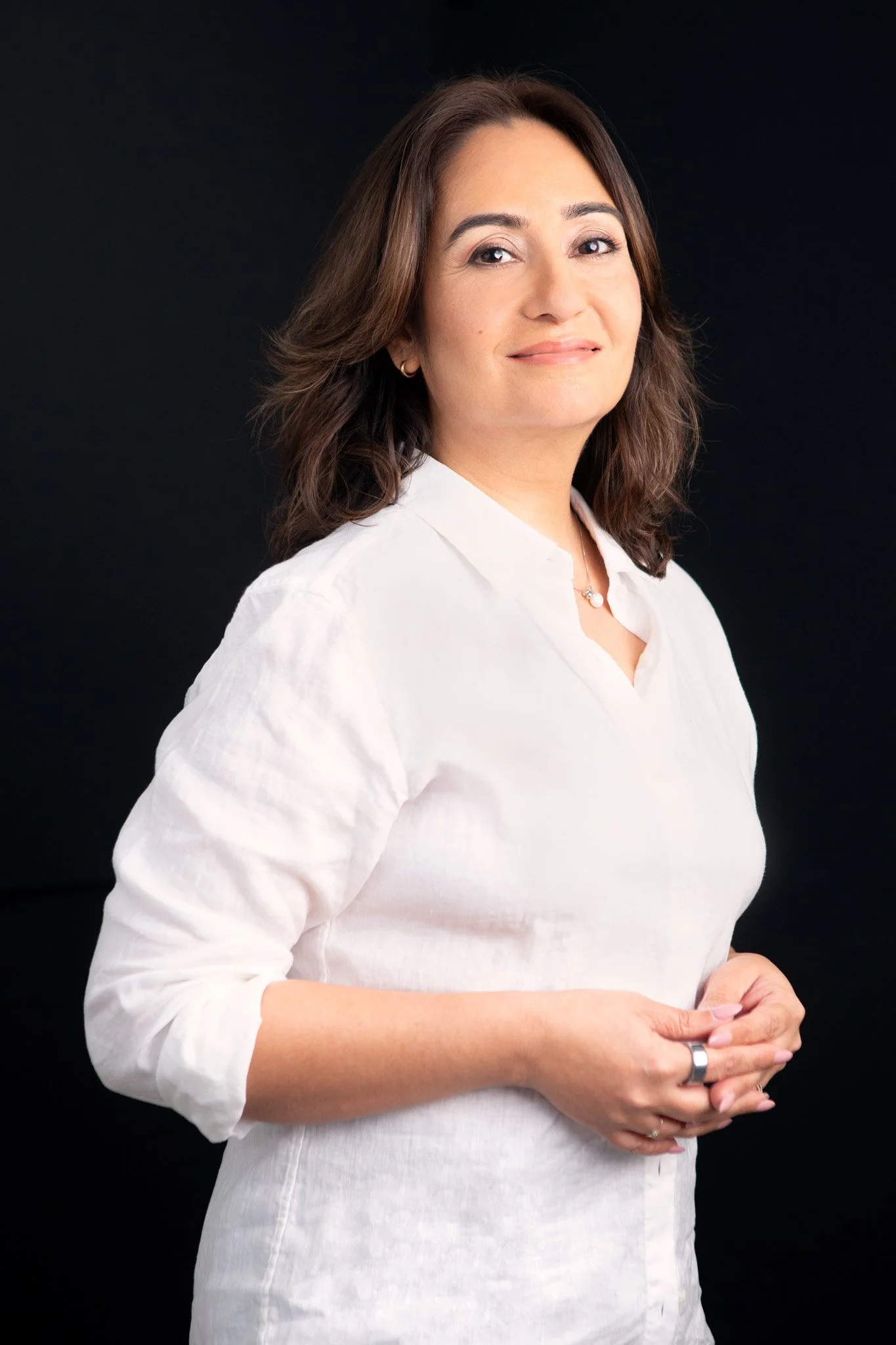 A woman with shoulder-length brown hair, wearing a white blouse, standing against a black background, with her hands gently clasped in front of her.
