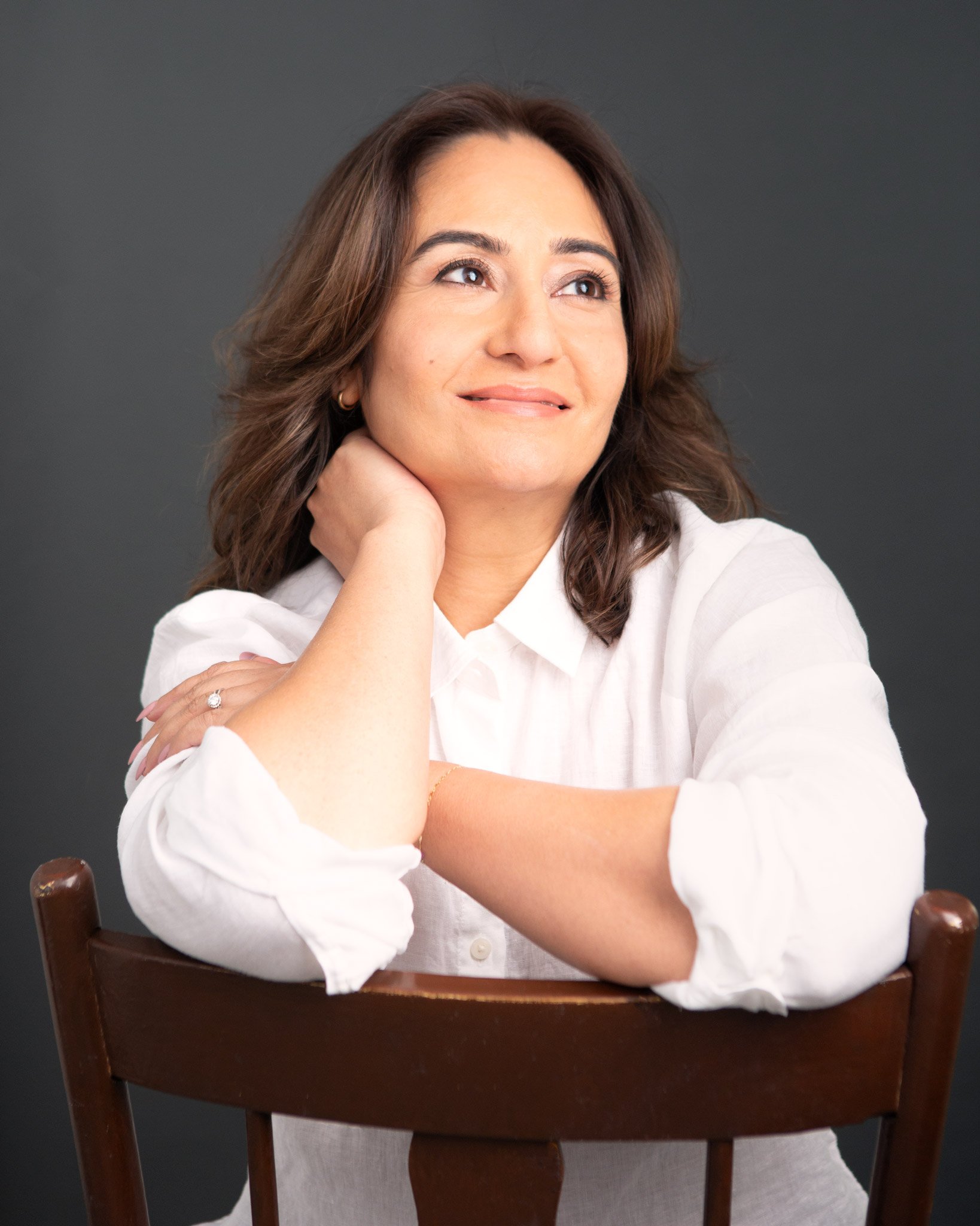 A woman with brown hair and light skin, wearing a white shirt, sitting behind a wooden chair, looking to her right with a slight smile, against a dark gray background.