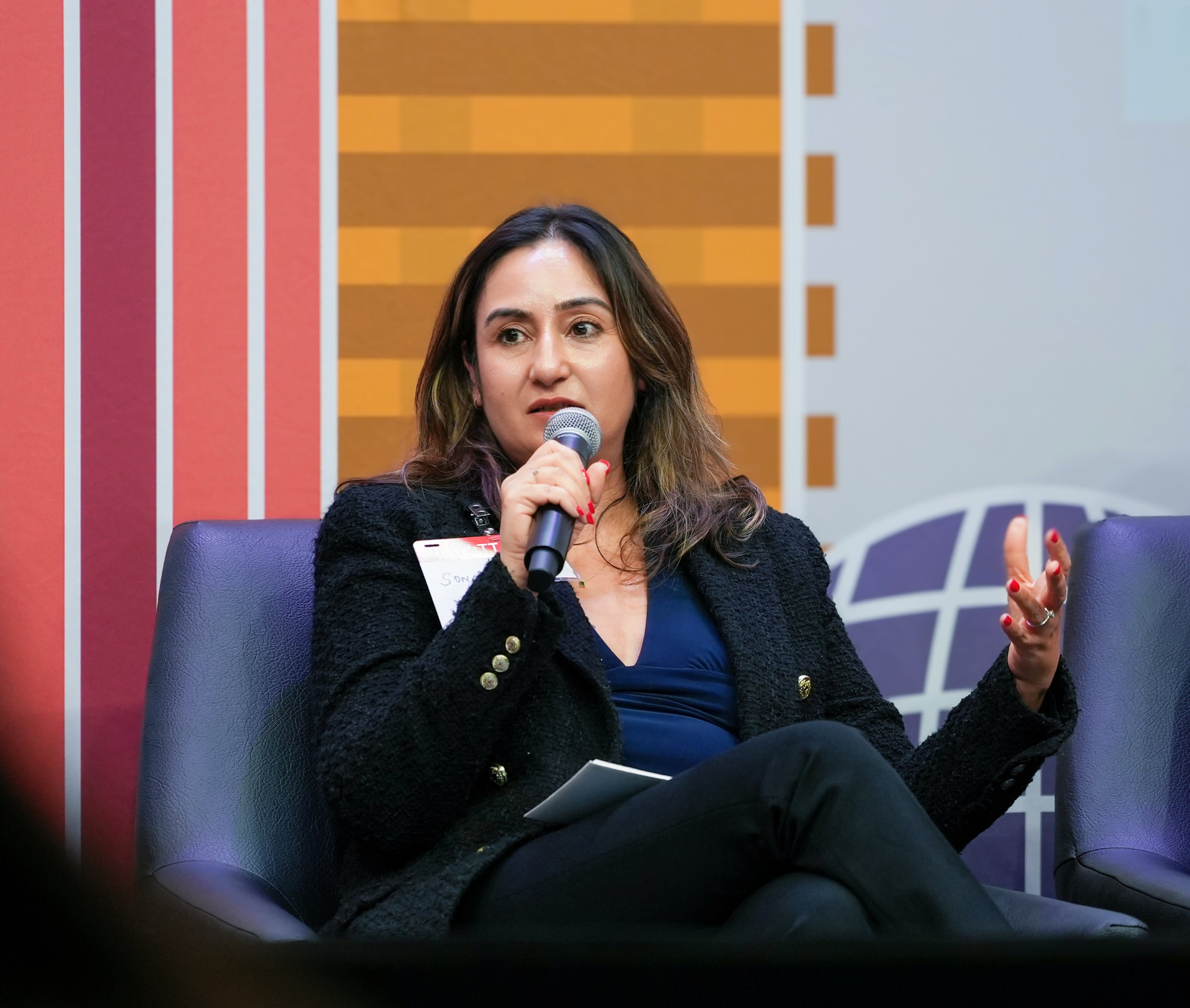 A woman is speaking into a microphone during an event or panel discussion. She has dark, wavy hair with lighter highlights and is wearing a black jacket with gold buttons and a blue top. She is sitting on a purple chair, holding a notepad, and has a name badge that says "Sonia." The background features a colorful abstract wall with red and orange stripes and a blue and white geometric design.