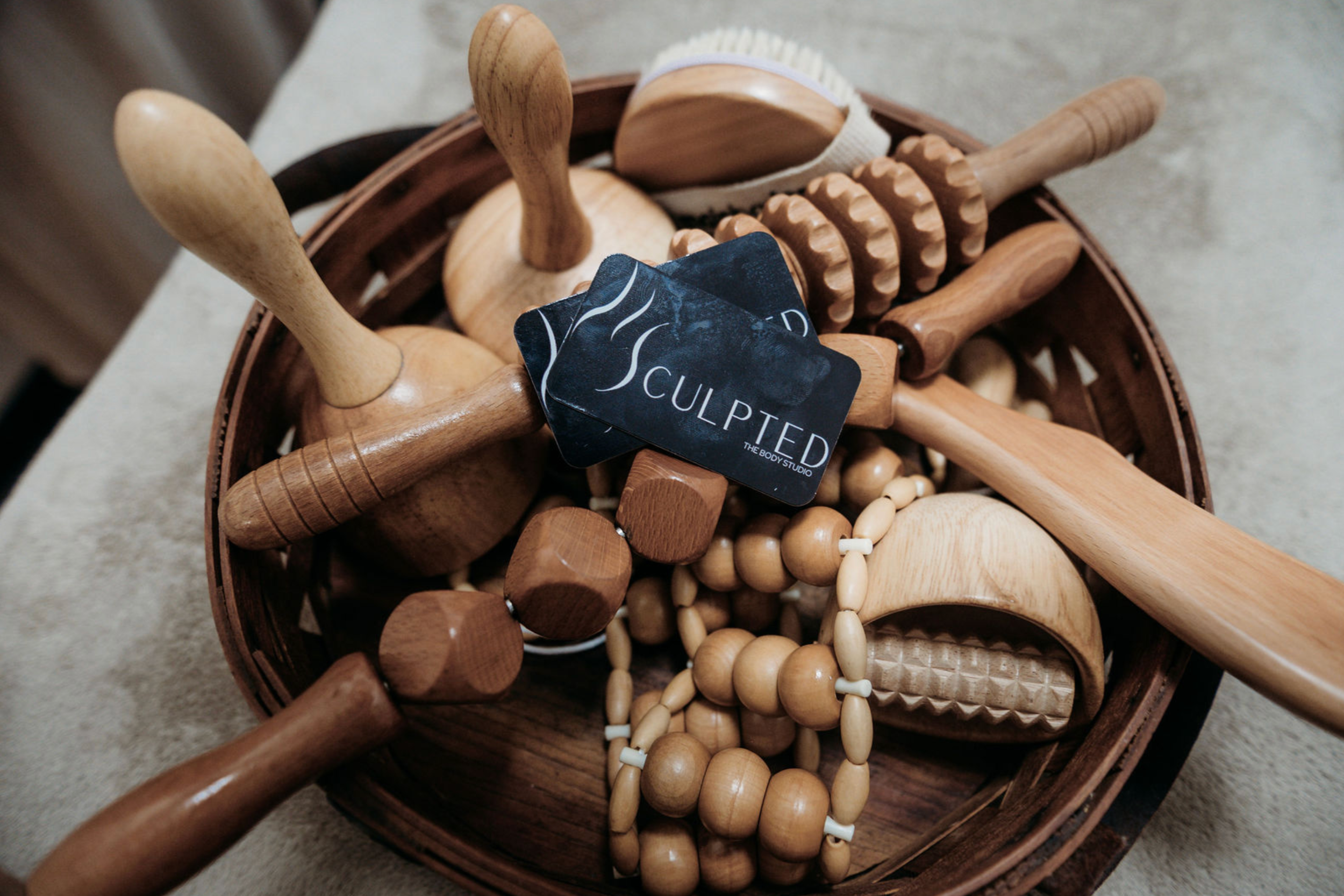 A basket containing wooden massage rollers and a tag that says 'sculpted' from the body studio, placed on a beige surface.