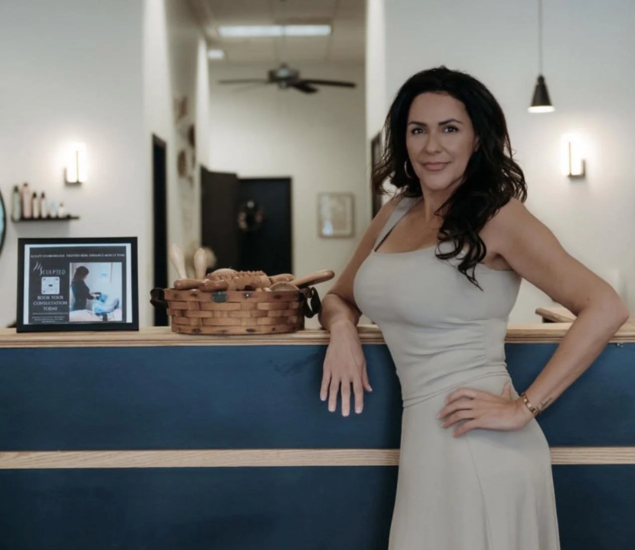 Woman in a beige dress leaning on a blue and light wood reception counter in a modern, well-lit interior.
