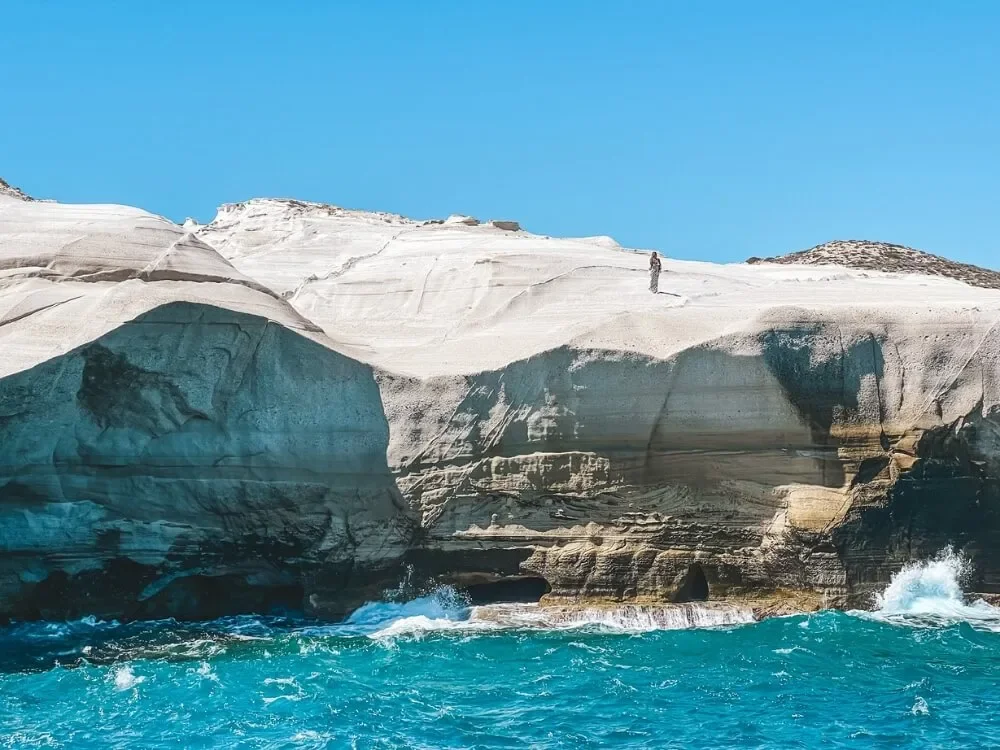 A rugged white coastal cliff with a person standing on top, overlooking the ocean with turquoise water and waves crashing against the rocks beneath.