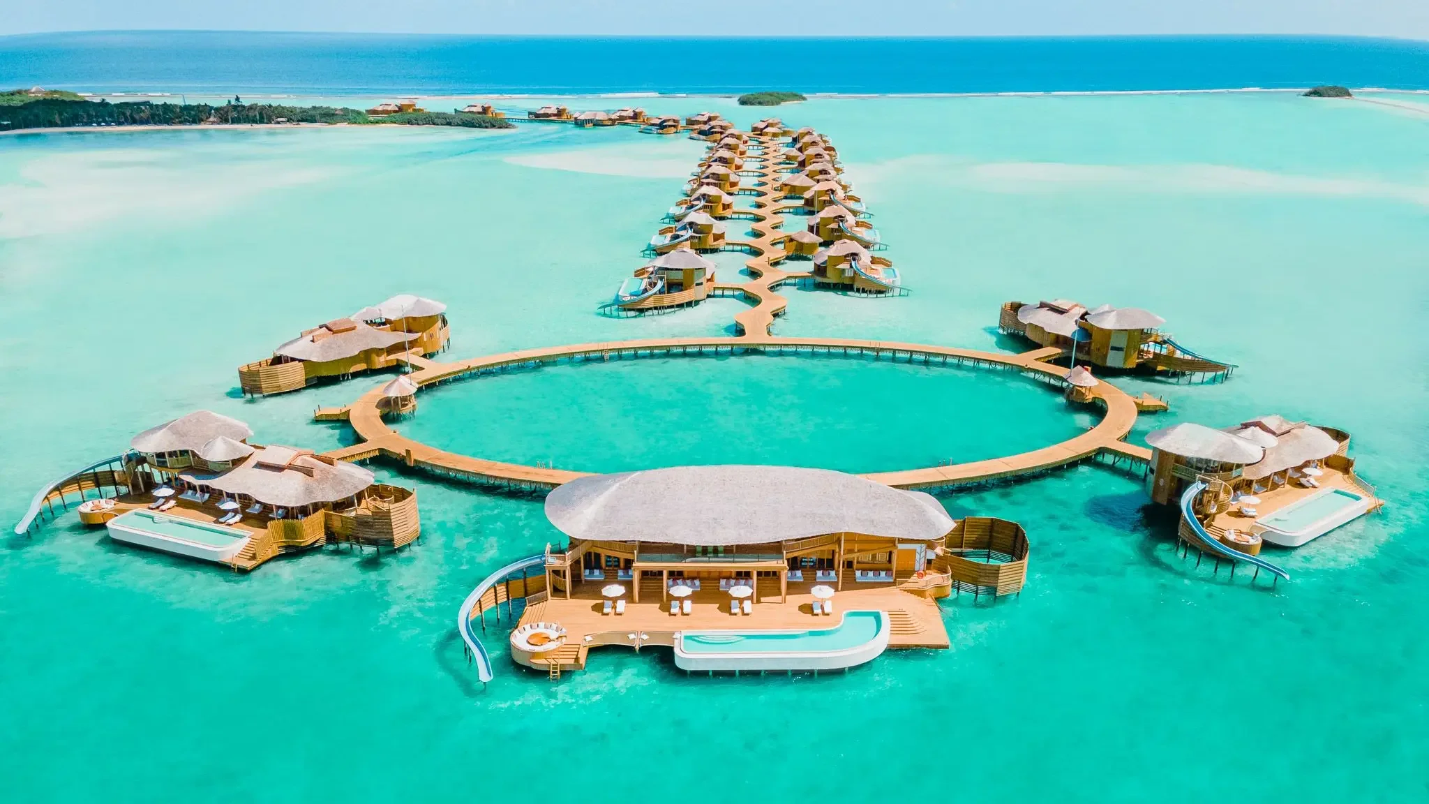 Overwater bungalows with thatched roofs connected by pathways over clear turquoise water at a tropical resort.