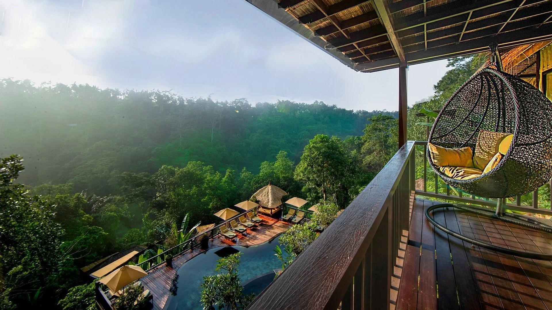 Balcony overlooking a lush, green jungle with poolside seating and umbrellas below, part of a tropical resort or hotel.