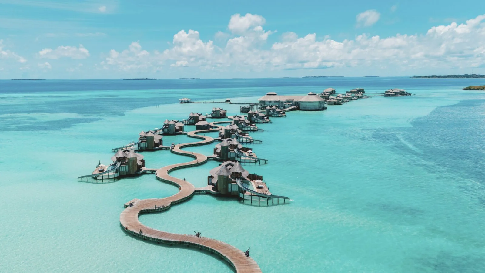 Overwater bungalows connected by a winding wooden boardwalk in clear blue ocean water under a partly cloudy sky.