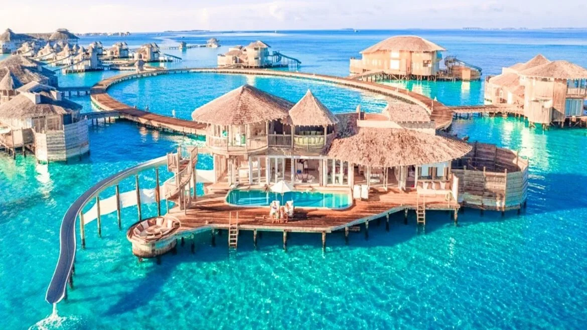 Overwater bungalow resort with interconnected wooden walkways, a water slide, and a pool, surrounded by clear blue ocean waters near a distant shoreline.