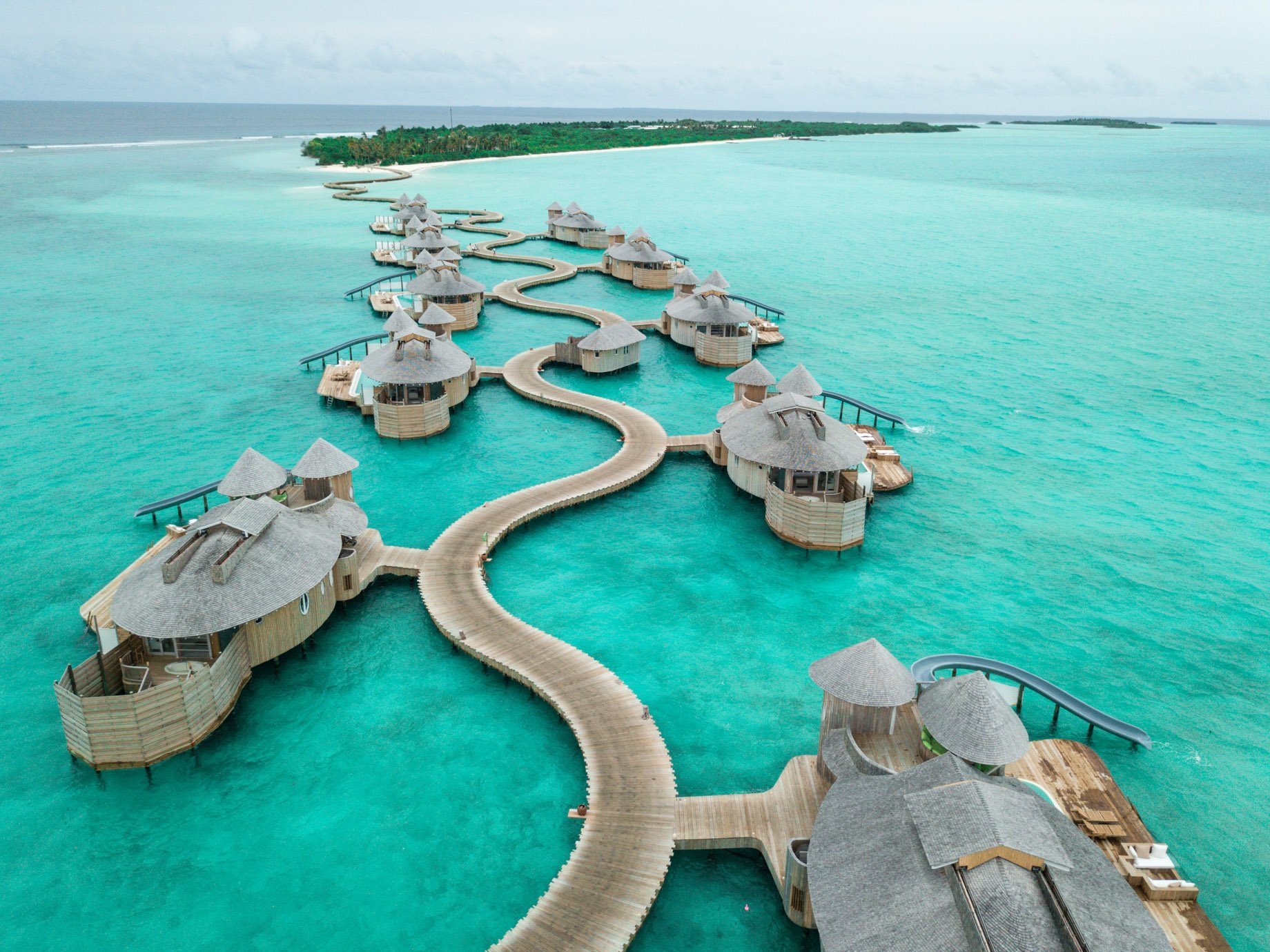 Overwater bungalows connected by a winding wooden walkway on a clear turquoise sea, with a lush green island in the distance.