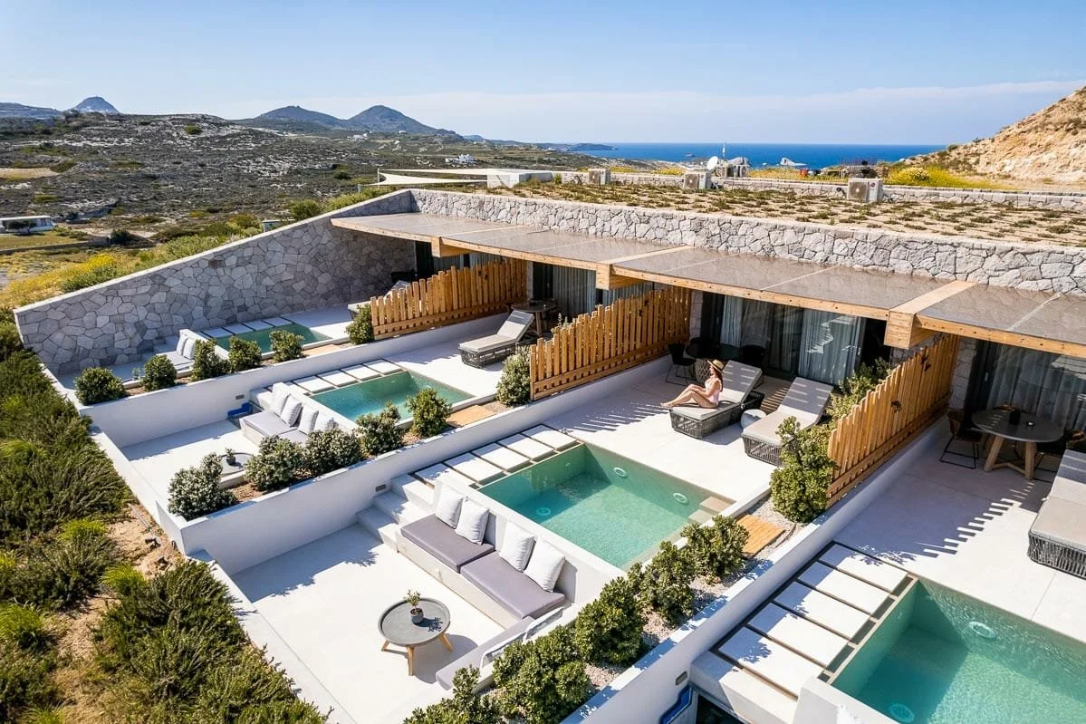 Modern outdoor pool area with two small pools, lounge chairs, and lush greenery, overlooking a hillside and the ocean in the distance.