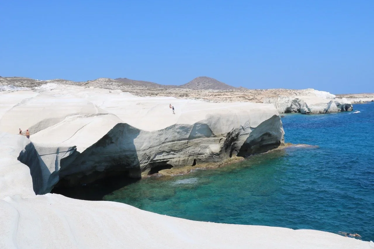 Coastal scene with white rocky cliffs and clear blue water, with a few people walking on the rocks under a bright blue sky.