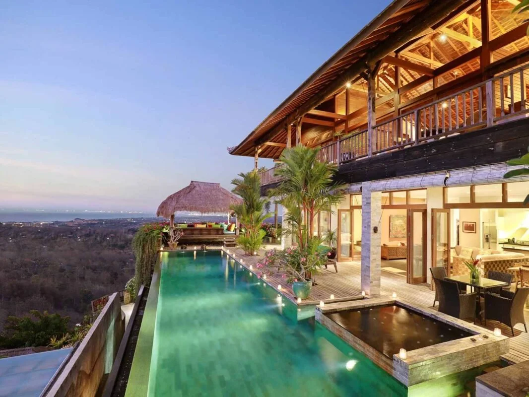 Luxury house with infinity pool overlooking a valley at dusk, with outdoor seating and tropical plants.